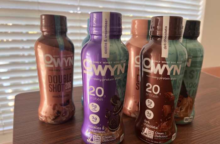 Five bottles OWYN Plant-Based Protein Shakes on a kitchen table, including the flavors Cookies & Creamless, Dark Chocolate and Double Shot Mocha Fudge